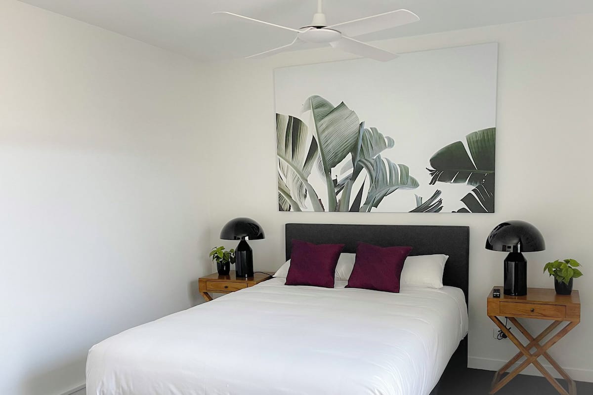 Un dormitorio moderno con una cama grande, dos mesitas de noche, un ventilador de techo y una obra de arte colgada en la pared.