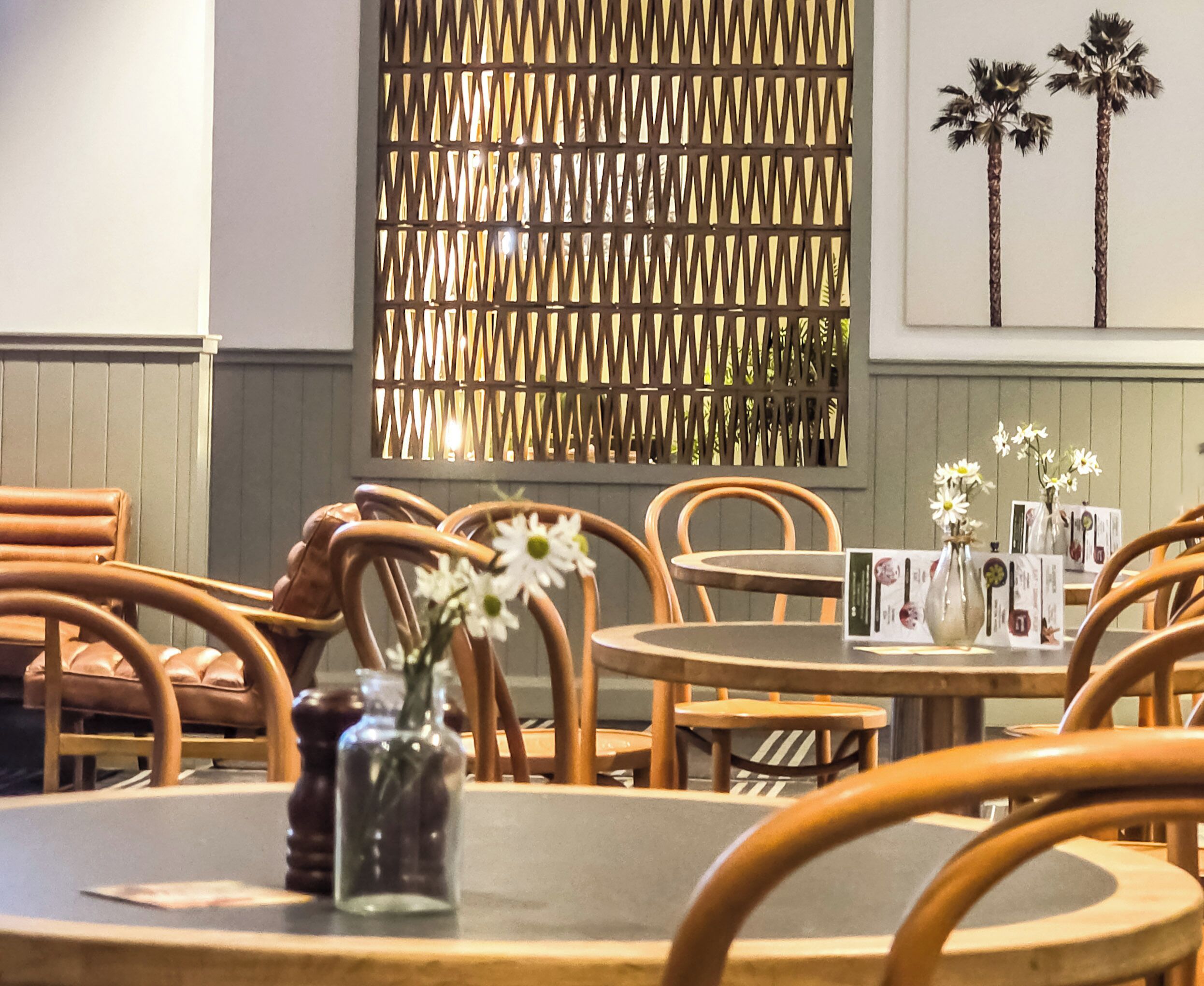 A restaurant interior with wooden chairs, round tables, a decorative wall, and a vase with flowers.