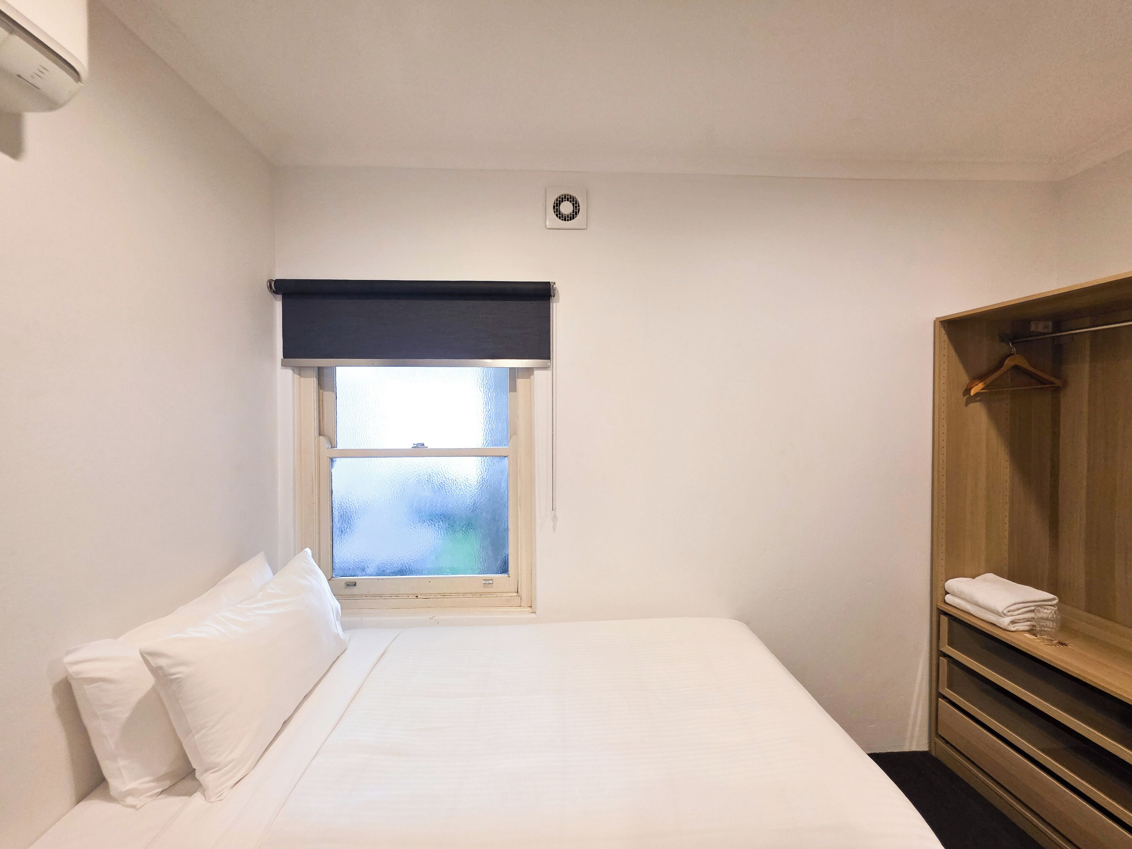 Une petite chambre à coucher, bien rangée, avec un lit, une fenêtre munie d’un store, une armoire et un thermostat fixé au mur.