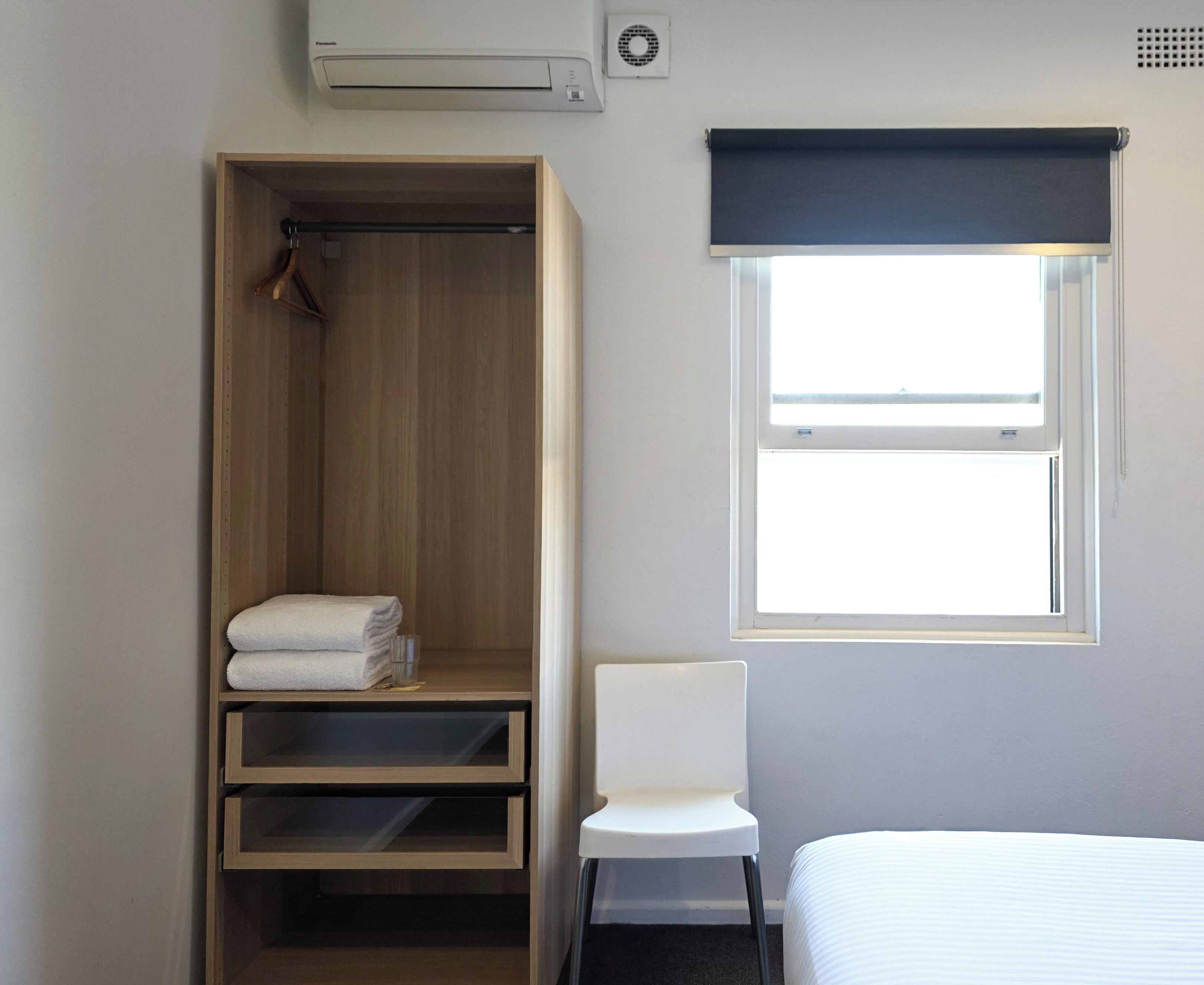 A hotel room with a wooden wardrobe, a white chair, a bed, and a window with a view.
