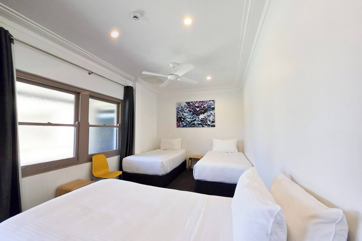 Habitación de hotel con dos camas, ventilador de techo, ventana con cortinas, una mesita y una obra de arte colgada en la pared.