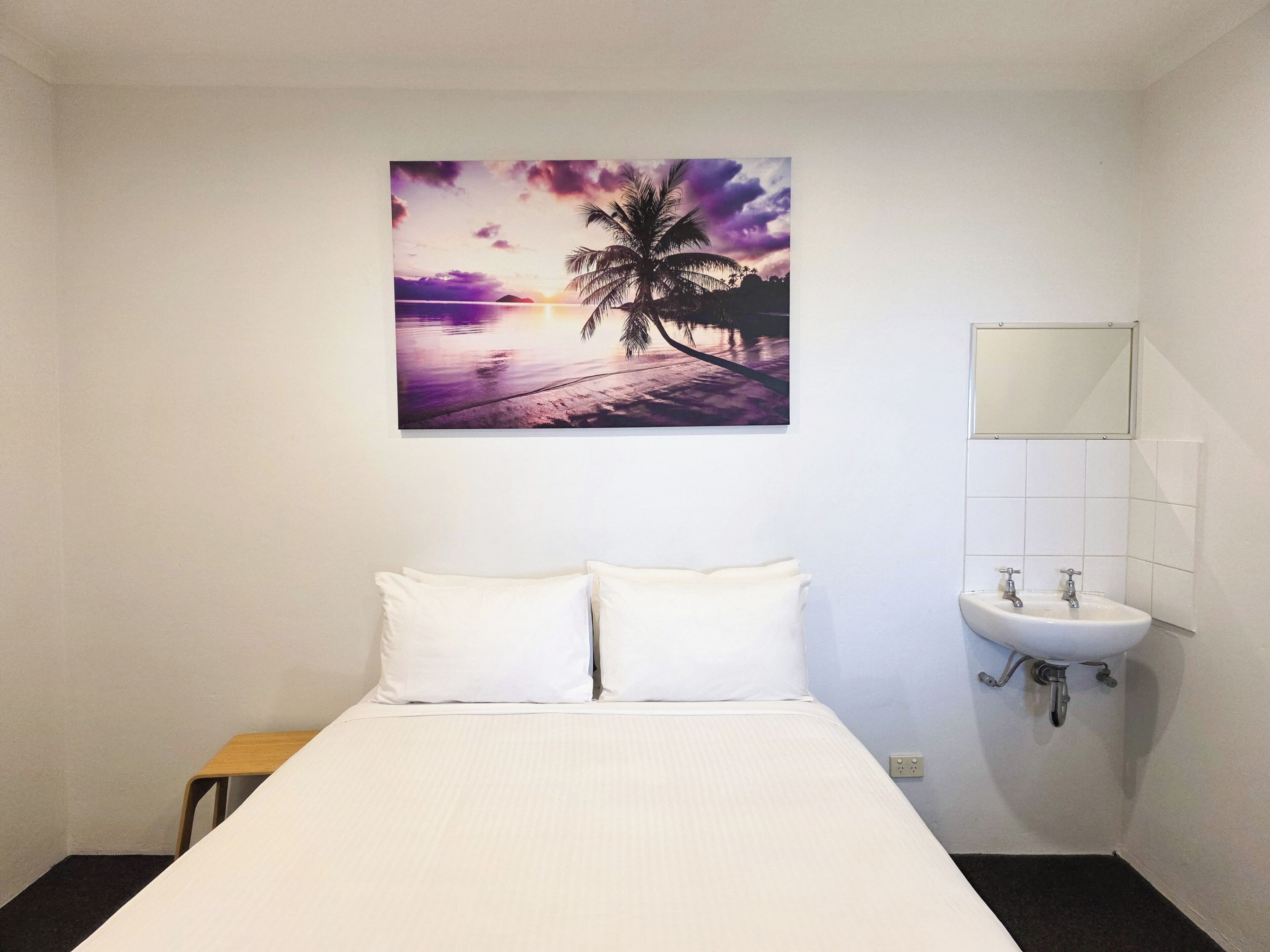 Une chambre d’hôtel avec un lit, une petite table, un lavabo et un tableau représentant un coucher de soleil sur une plage.