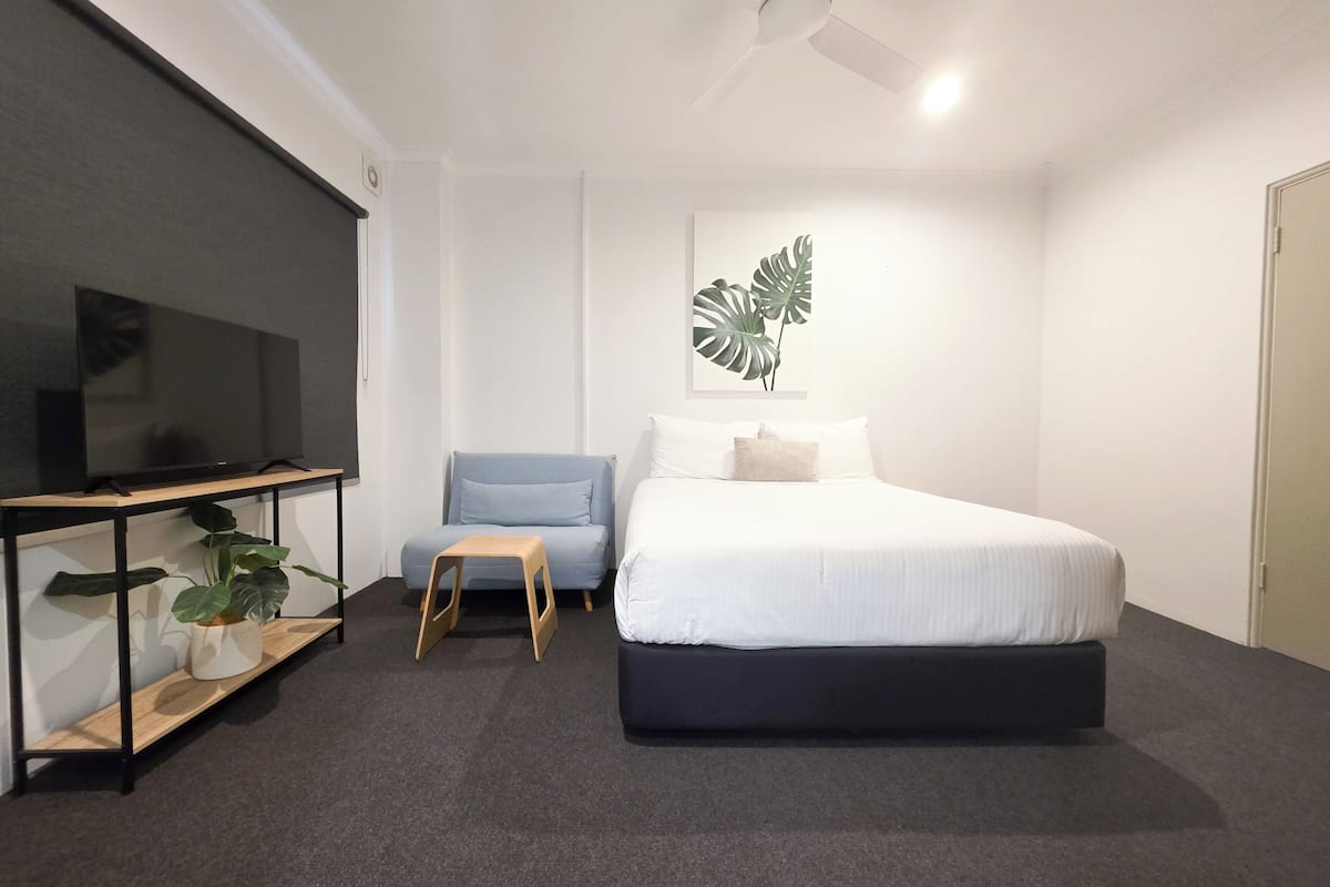 Un dormitorio con una cama, un televisor sobre un mueble bajo, una silla y un ventilador de techo.