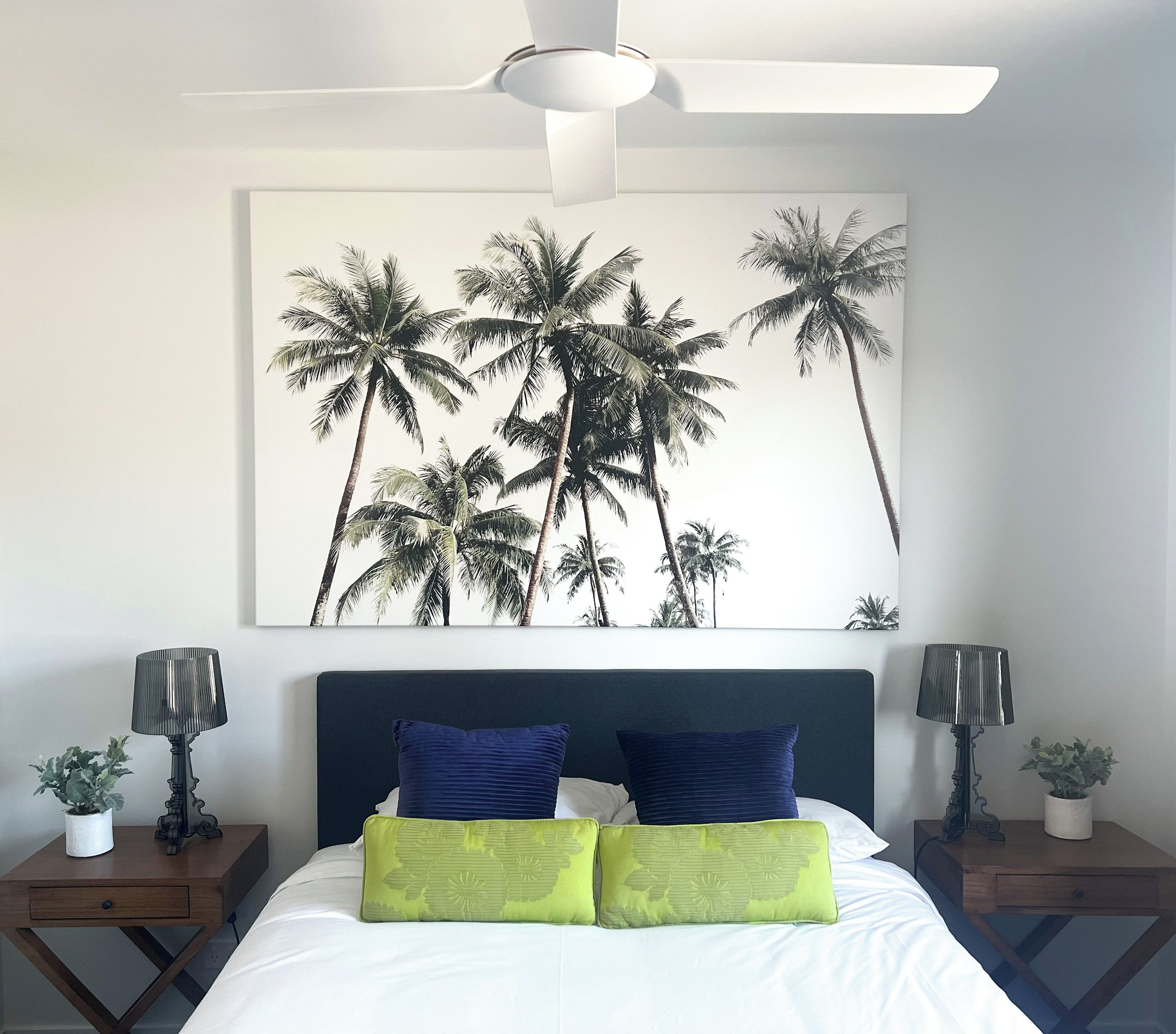 A bedroom with a bed, two nightstands, a ceiling fan, and a large wall art of palm trees.