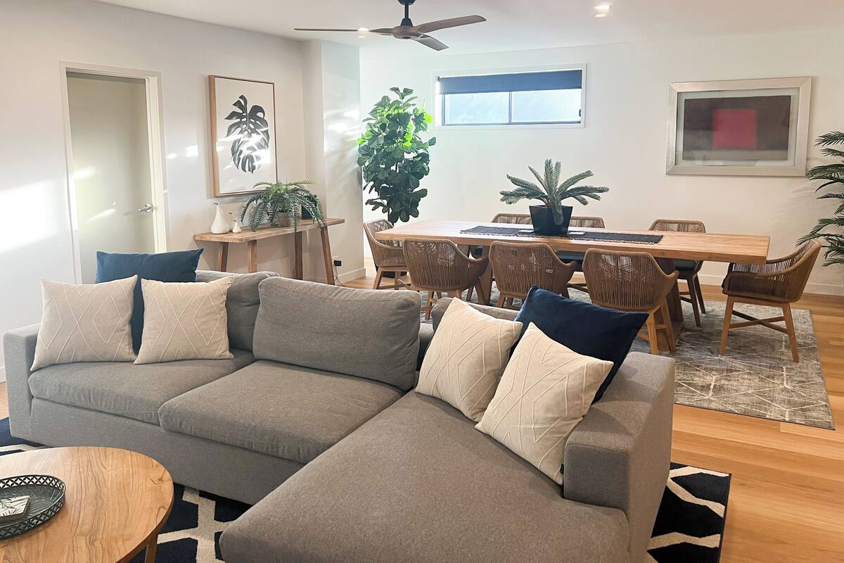 Una sala de estar moderna con un sofá modular, una mesa de comedor y sillas. Hay un ventilador de techo, un ventanal grande y plantas en macetas.