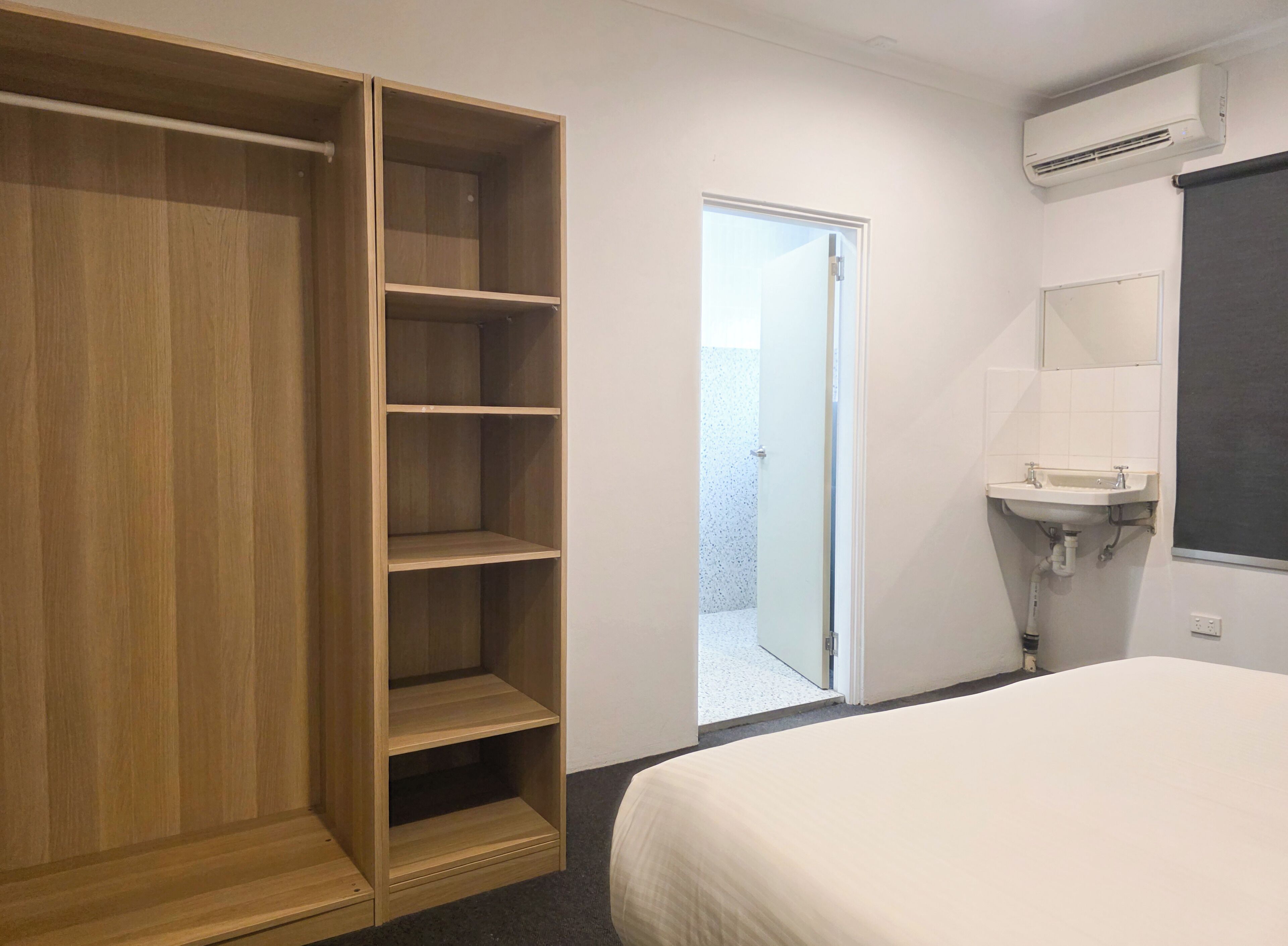 A bedroom with a wooden wardrobe, a white bed, a bathroom with a sink and mirror, and an air conditioning unit.