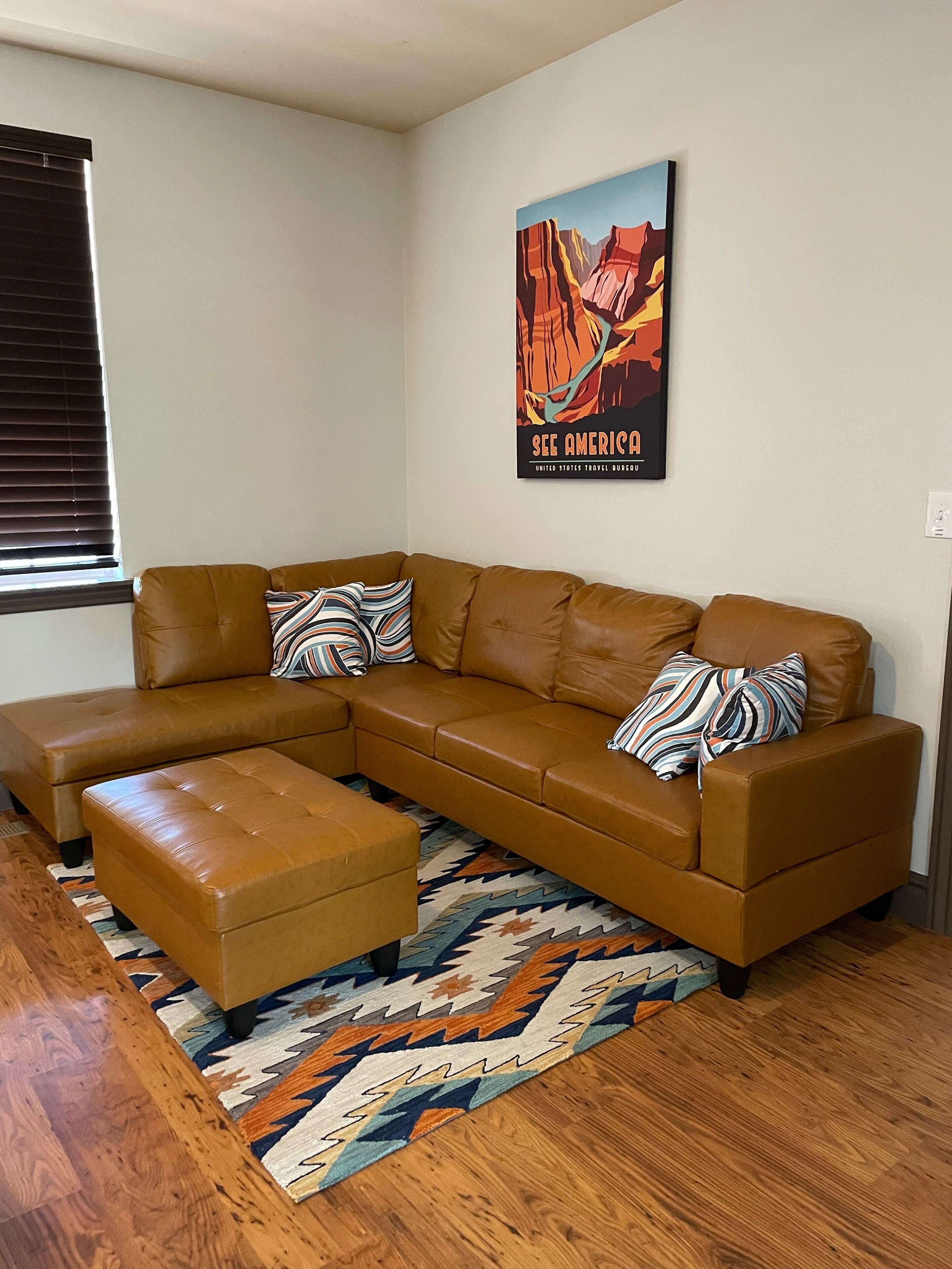 A modern living room with a brown leather sectional sofa, a matching ottoman, and a colorful rug.