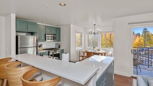 A modern kitchen with a central island, teal cabinets, stainless steel appliances, and a dining area with a view of autumn trees.