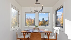 A dining room with a wooden table, four chairs, and a chandelier. There are large windows with a view of trees and mountains.