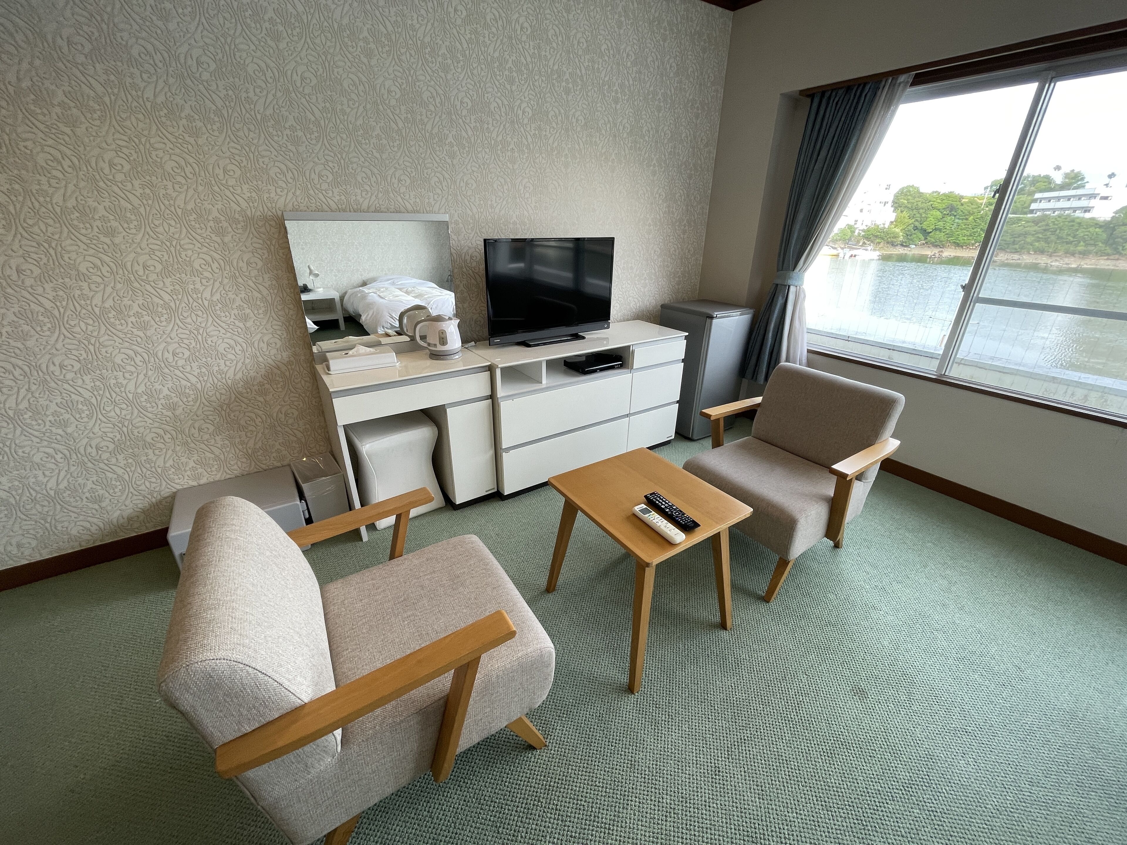 A hotel room with a bed, a TV, a desk, a chair, a small table, and a window with a view of a body of water.