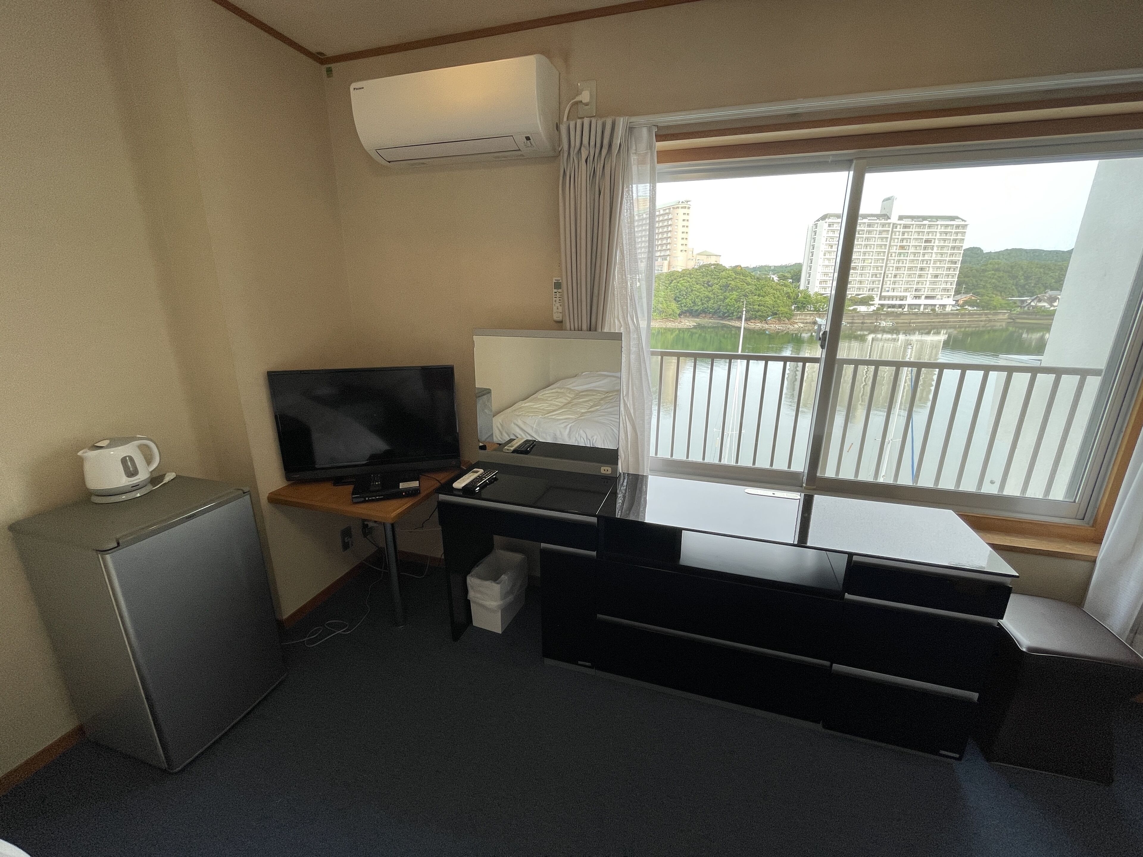 Et lille hotelværelse med seng, skrivebord med computer, fjernsyn, minikøleskab og et vindue med udsigt til bygninger og grønne områder.