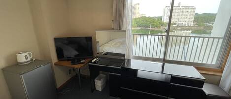 Et lille hotelværelse med seng, skrivebord med computer, fjernsyn, minikøleskab og et vindue med udsigt til bygninger og grønne områder.