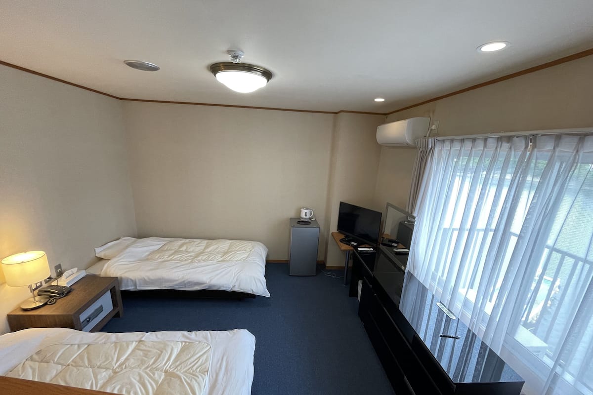 A hotel room with two beds, a desk with a TV, a coffee machine, and a window with sheer curtains.