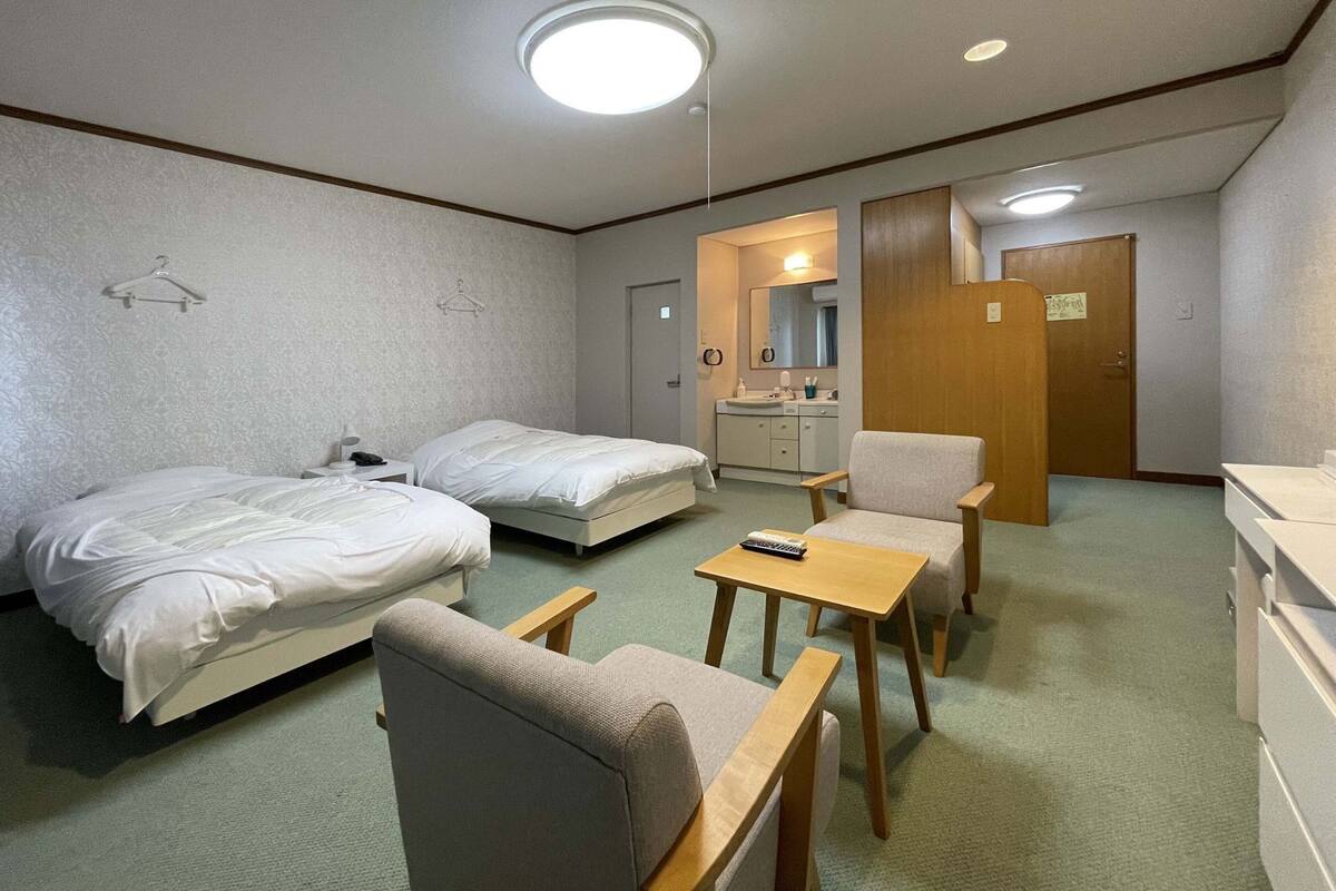 A hotel room with two beds, a small table, a chair, a desk, a mirror, a sink, and a door.