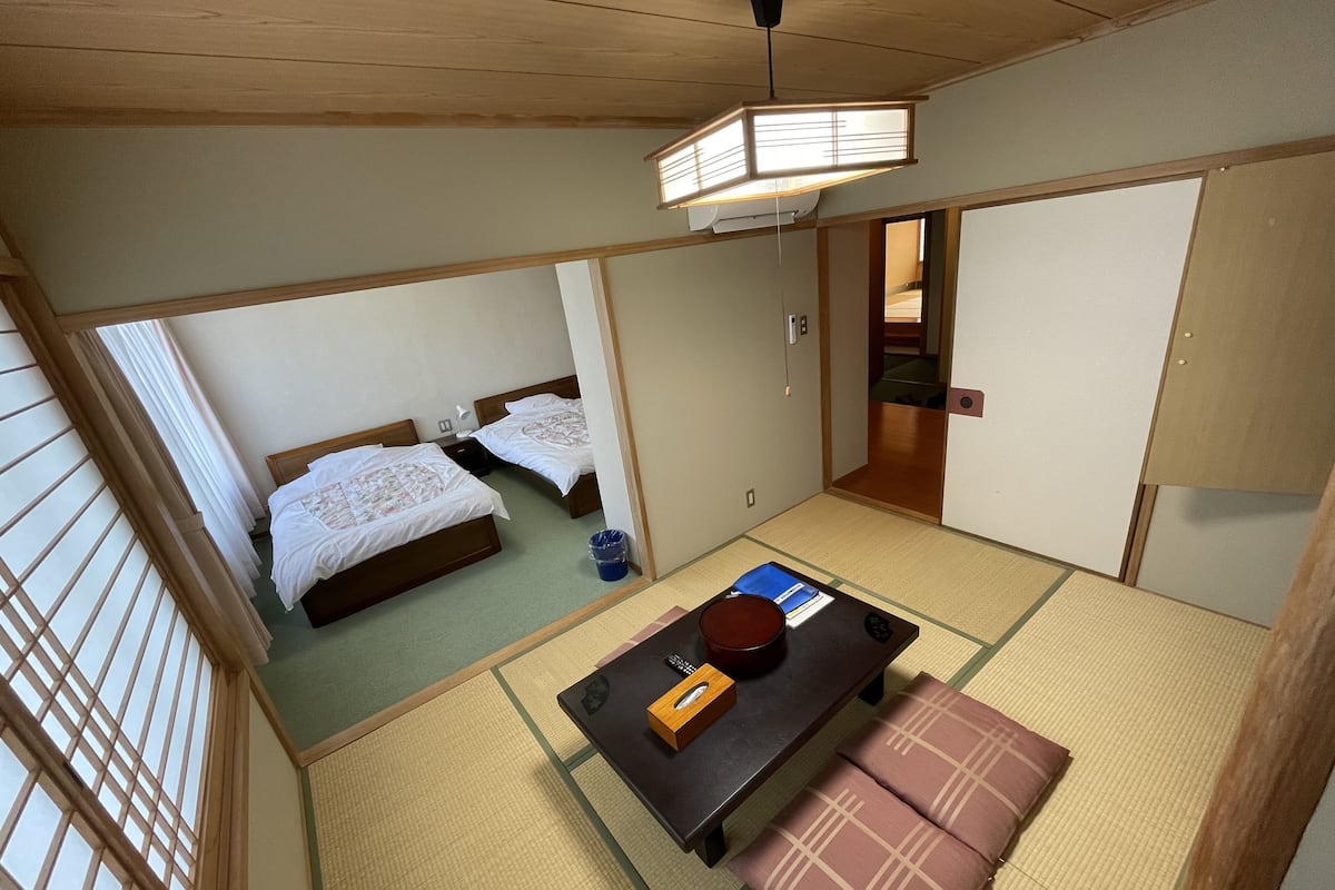 A traditional Japanese-style room with tatami flooring, two beds, a table with a remote control, and a hanging light fixture.