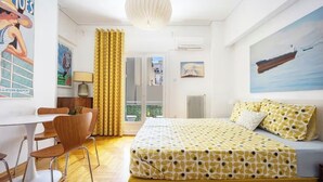 Quarto de hotel com cama, escrivaninha e cadeiras, janela com cortinas e um quadro na parede.