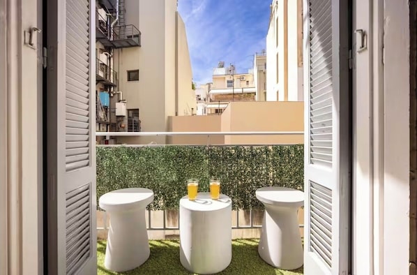 Ein Balkon mit drei weißen Hockstühlen, zwei Orangensaftgläsern und einer Hecke im Hintergrund.