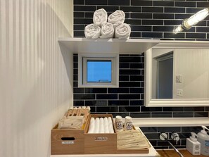 A bathroom with a black and white tiled backsplash, a small window, and a shelf with towels and toiletries.