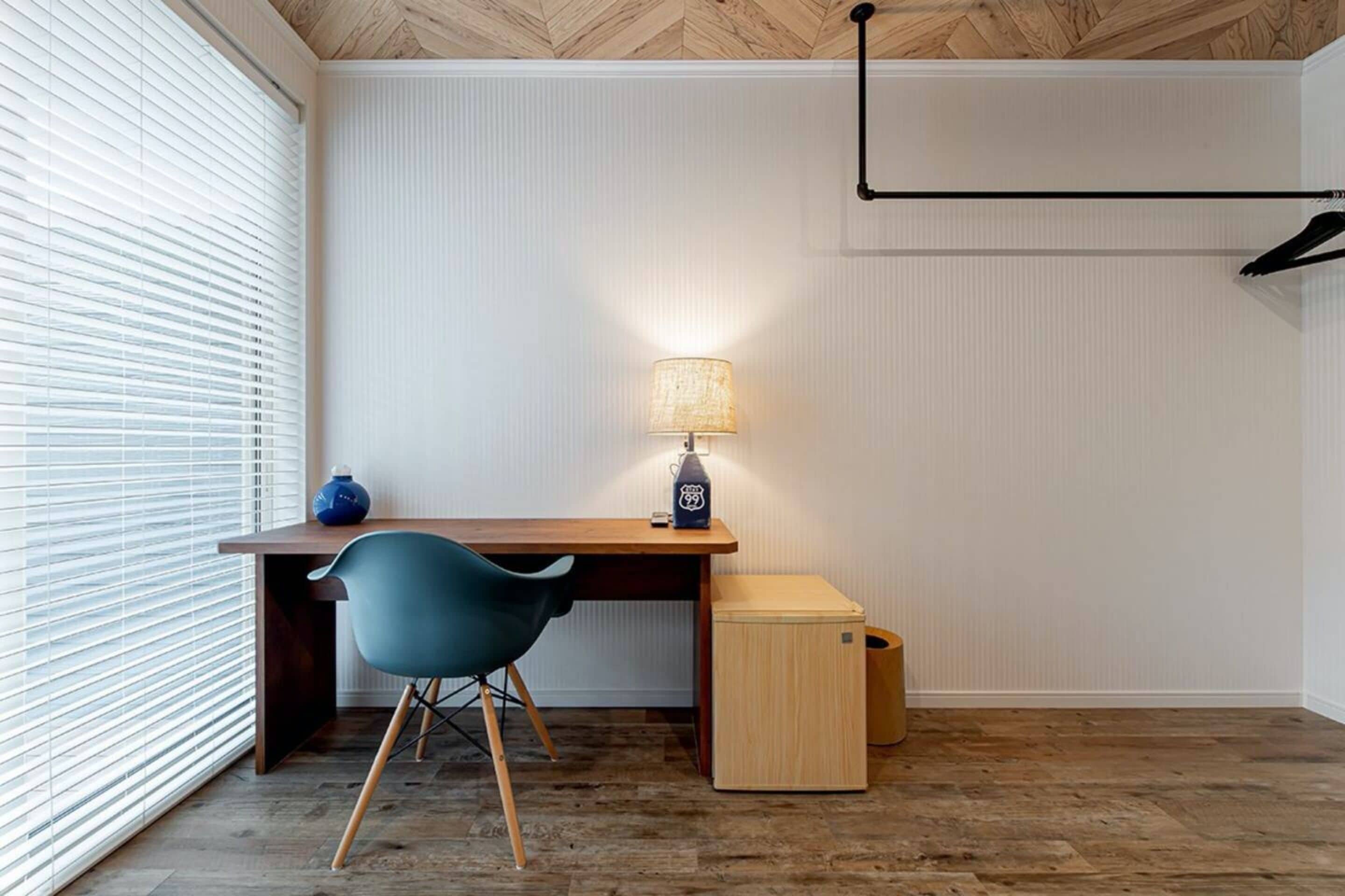 A room with a wooden desk, a blue chair, a lamp, and a small wooden cabinet.