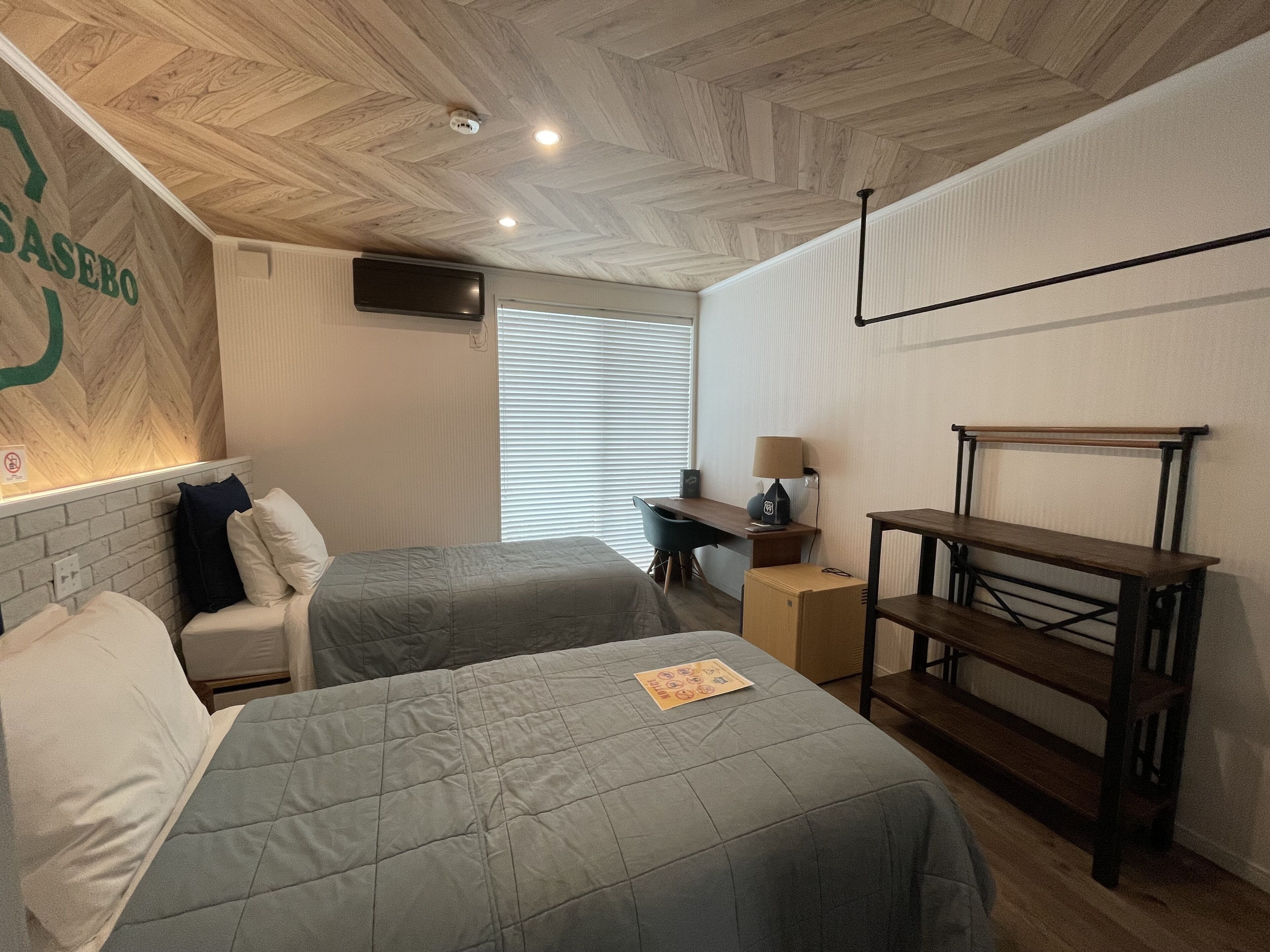 A hotel room with two beds, a desk, a chair, a lamp, and a bench.