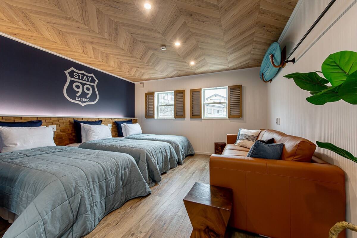Habitación de hotel con techo de madera, dos camas con ropa de cama gris, un sofá marrón y una planta verde grande.