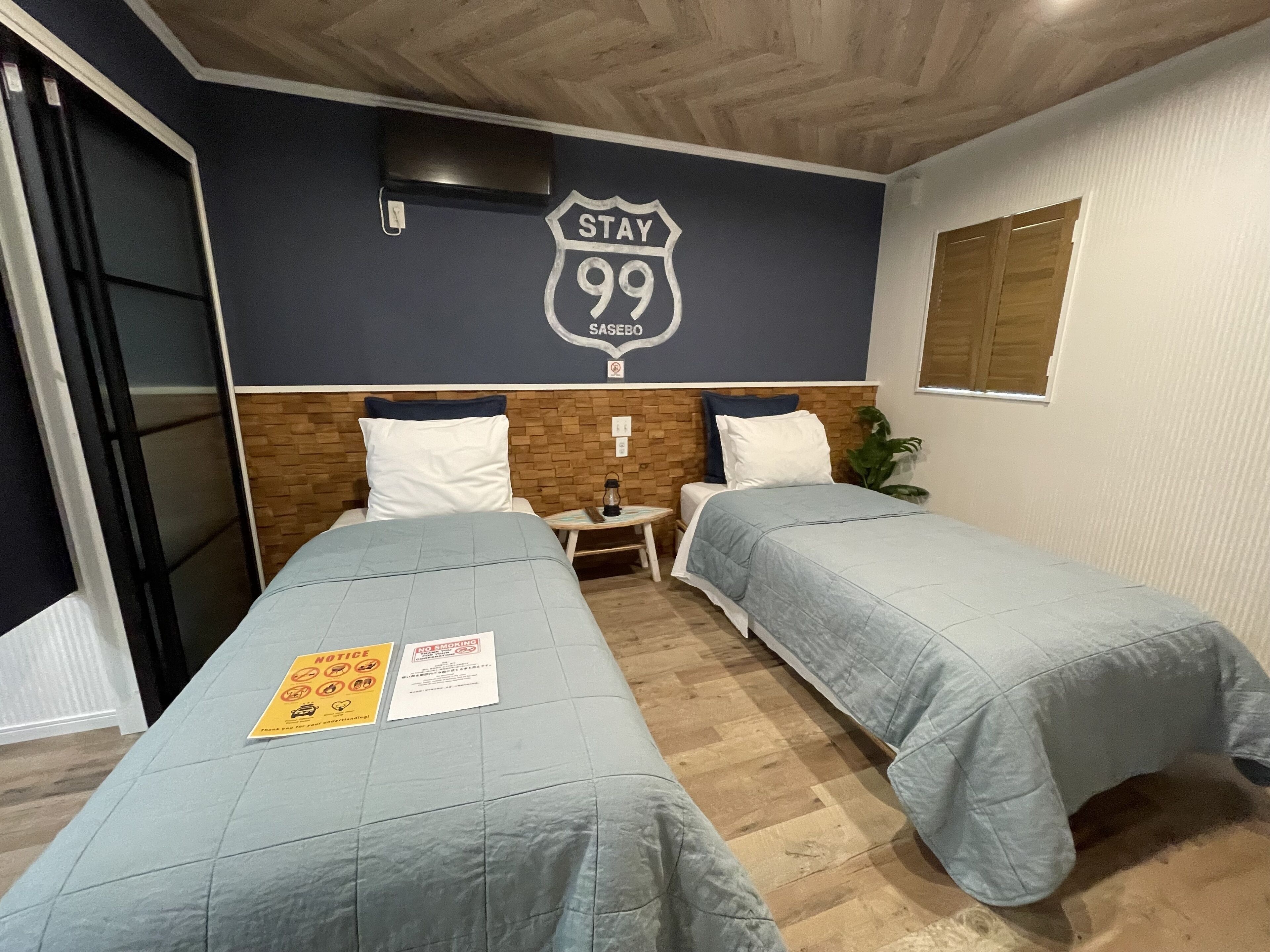 Habitació d'hotel amb dos llits, un panell de fusta al capçal, un televisor fixat a la paret i un cartell de 'Stay 99 Sasebo'.