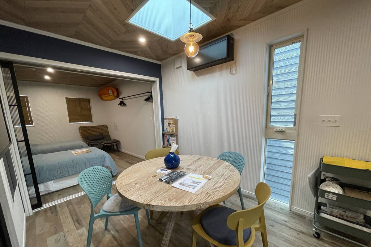 Un espacio habitable pequeño con una mesa redonda de madera, dos sillas, un televisor montado en la pared y una pequeña zona de kitchenette.