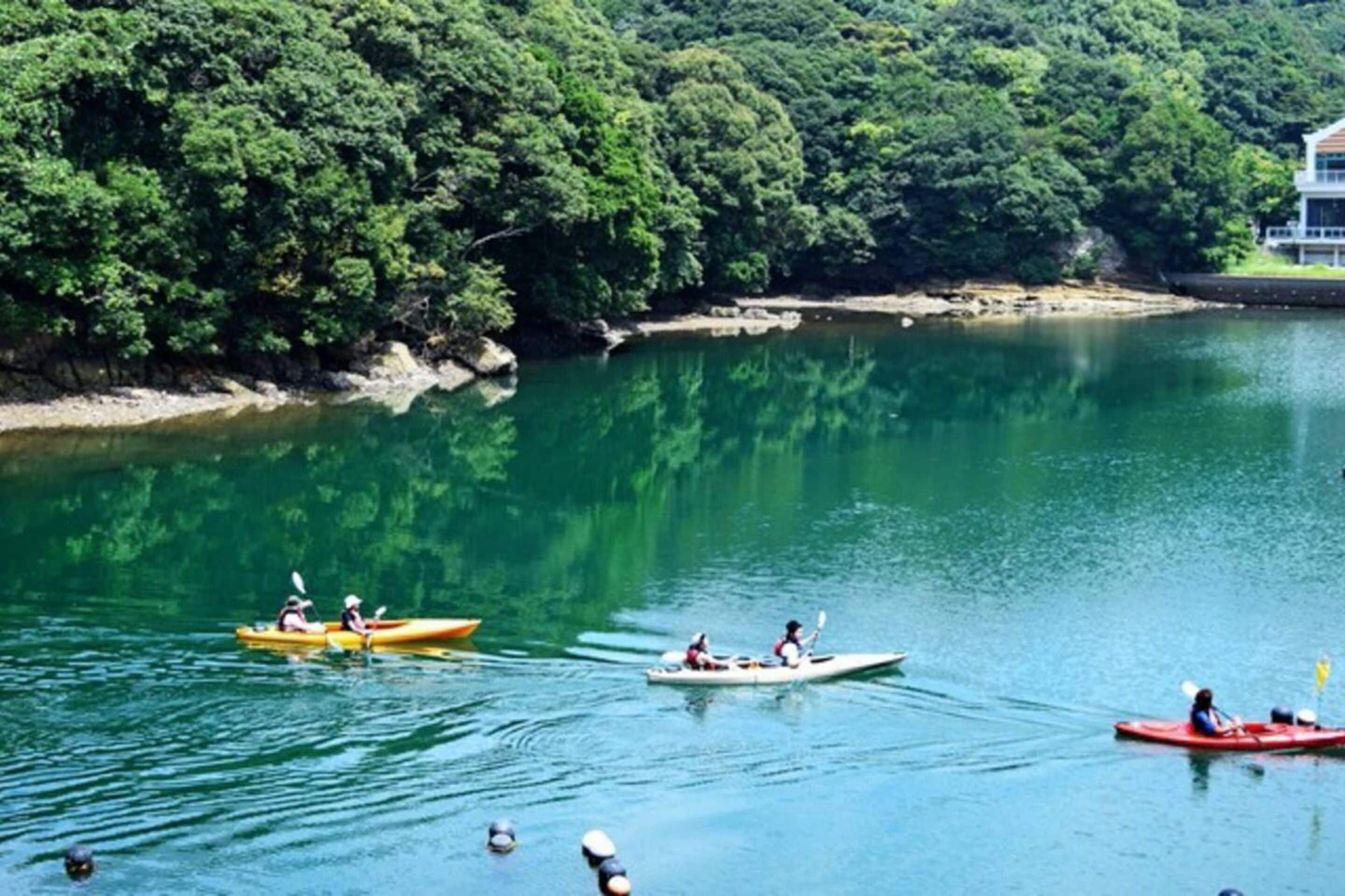 A calm lake with kayaks and canoes, surrounded by dense greenery and a building on the shore.