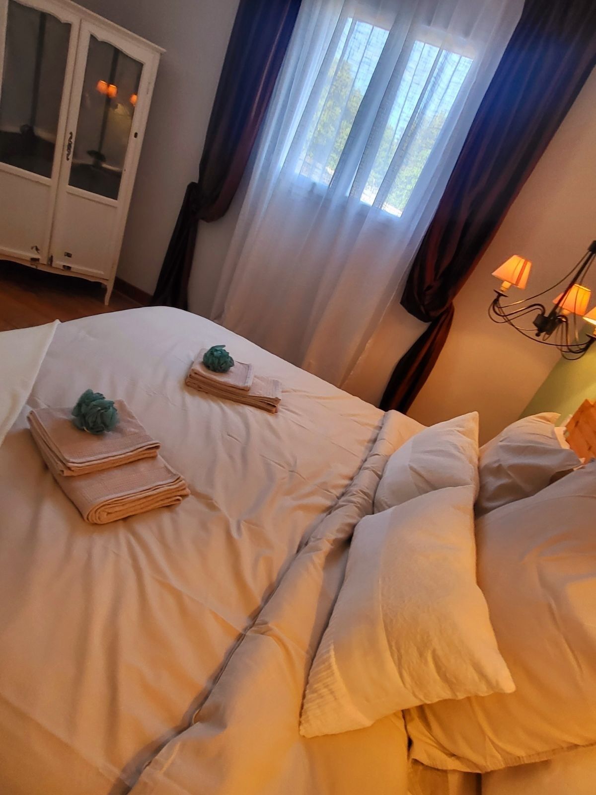 A neatly made bed with folded towels and a small plant, a white cabinet with glass doors, and a window with curtains.