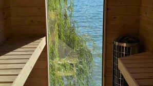 A wooden cabin with a large window overlooking a lake and mountains. Inside, there are wooden benches and a trash bin. - Private Lakefront Beach House off Naramata Bench with sauna and hot tub (Penticton)