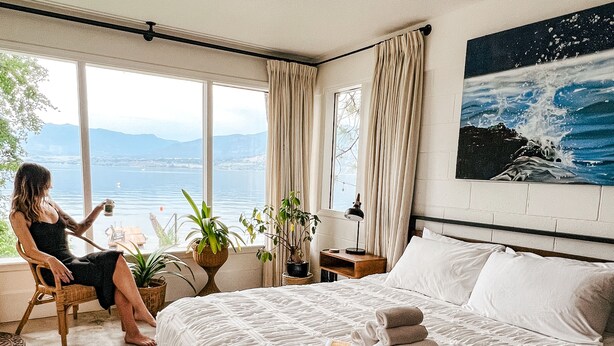 A bedroom with a large bed, a view of the sea, and a person sitting by the window. - Private Lakefront Beach House off Naramata Bench with sauna and hot tub (Penticton)