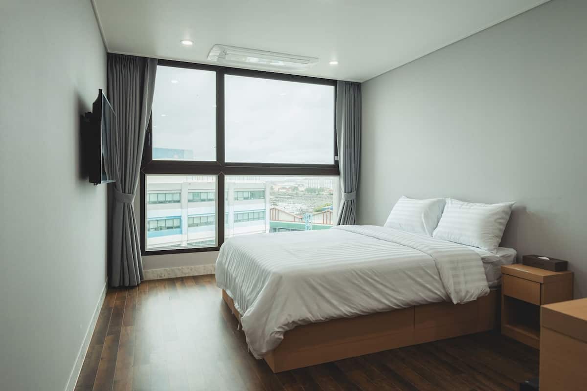 A modern bedroom with a large window, a bed, a nightstand, and a TV mounted on the wall.
