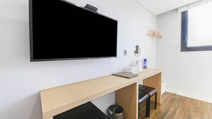 Una habitación de hotel moderna con un televisor de pantalla plana grande montado en la pared, un escritorio de madera con una computadora portátil, un cesto de basura pequeño y una ventana con vistas al cielo.