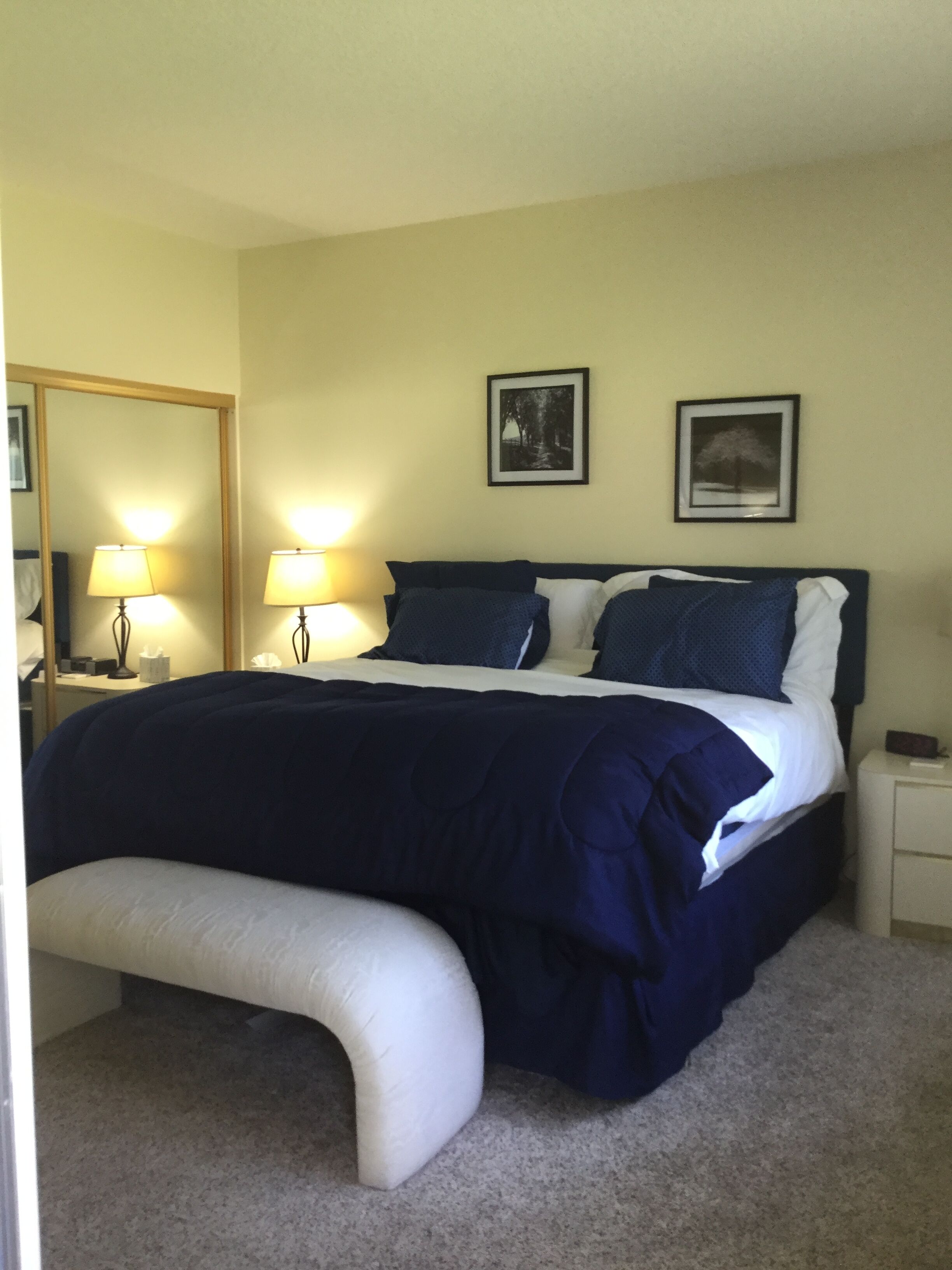 A bedroom with a bed, two bedside lamps, a nightstand, a mirror, and two framed pictures on the wall.