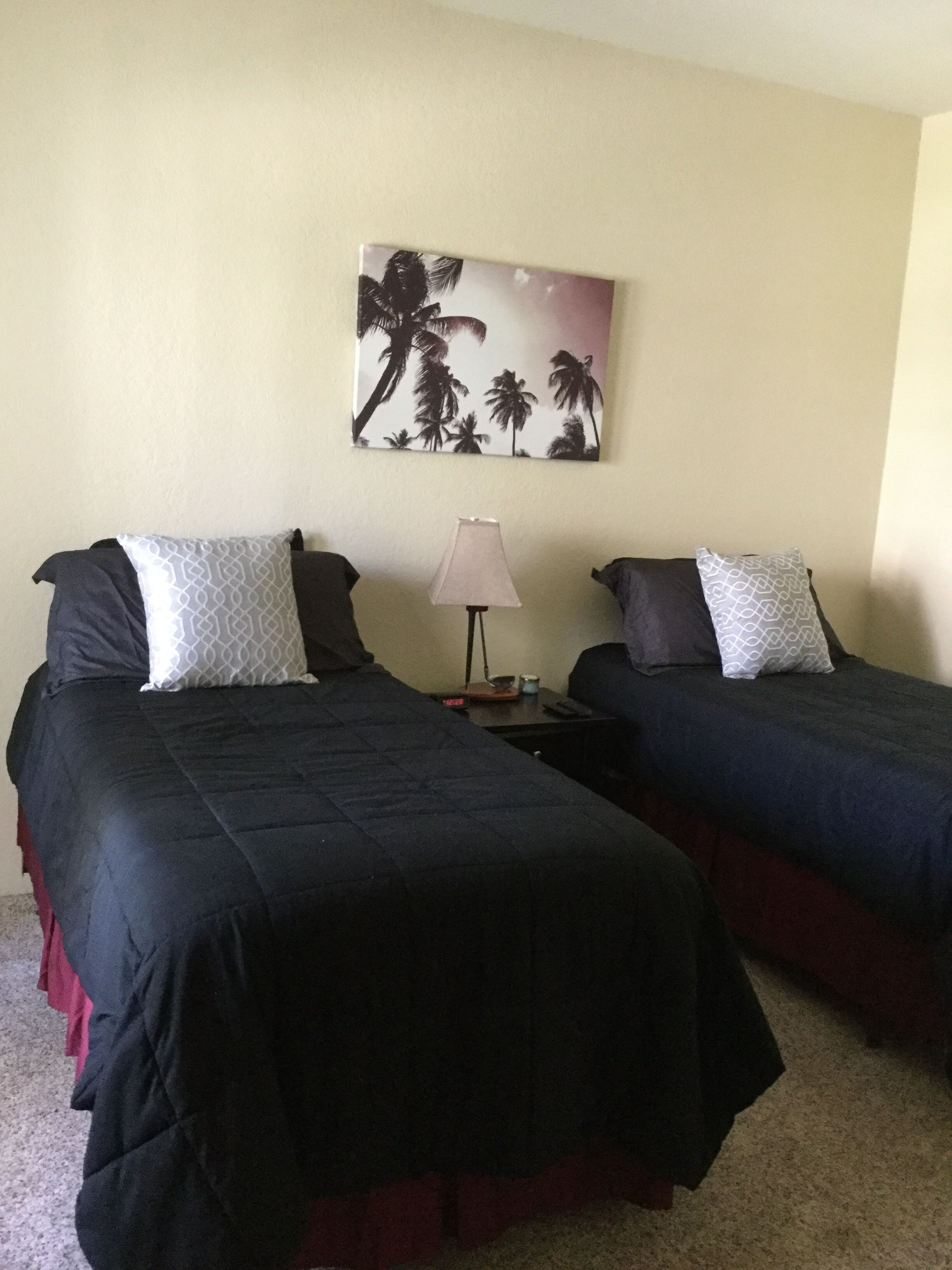 A hotel room with two beds, a nightstand with a lamp, and a painting of palm trees on the wall.
