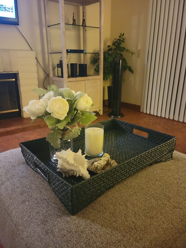 A room with a black woven tray on a carpet, containing a vase with white flowers, a candle, and decorative items.