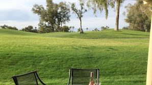 A covered patio with a round table and chairs, a bottle of wine, and a glass. The patio overlooks a grassy area with trees and hills in the background. - Desert Falls Country Club (Palm Desert)
