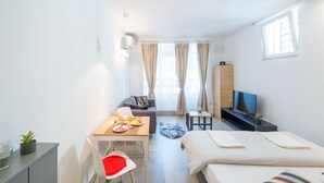 A modern hotel room with a bed, a desk with a chair, a sofa, a TV, and a window with curtains. - Apartment Kantun (Dubrovnik)