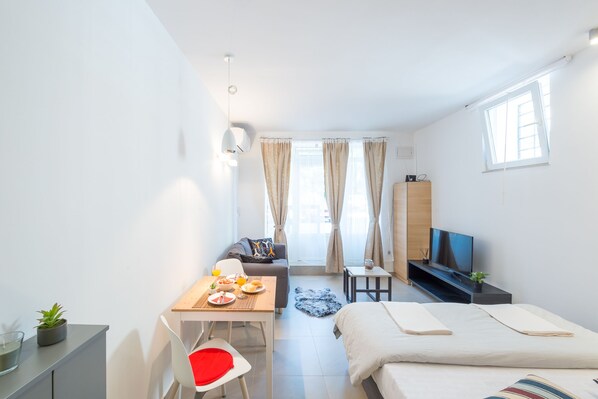 A modern hotel room with a bed, a desk with a chair, a sofa, a TV, and a window with curtains. - Apartment Kantun (Dubrovnik)