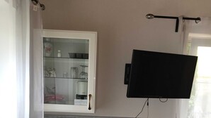 Ett litet rum med ett skrivbord, en stol, en tv som är monterad på väggen, ett skåp med glasdörrar och ett fönster med gardiner.