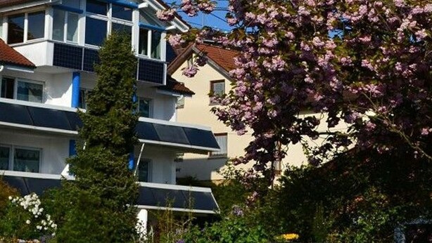 En flervåningsbyggnad med bostäder och balkonger, omgiven av grönska och blommande träd.
