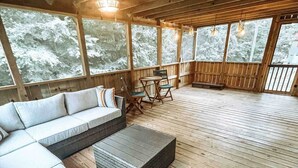 Een houten terras met een bank en stoelen, grote ramen met uitzicht op sneeuw en een hangende lamp.
