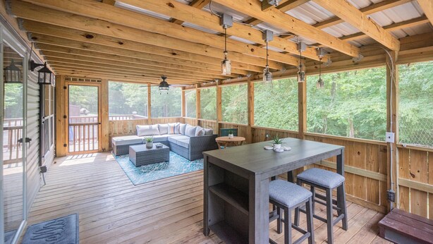 Een overdekt terras buiten, met houten balken, een bank, een tafel met krukken en uitzicht op het omliggende bos.