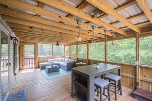 Een overdekt terras buiten, met houten balken, een bank, een tafel met krukken en uitzicht op het omliggende bos.