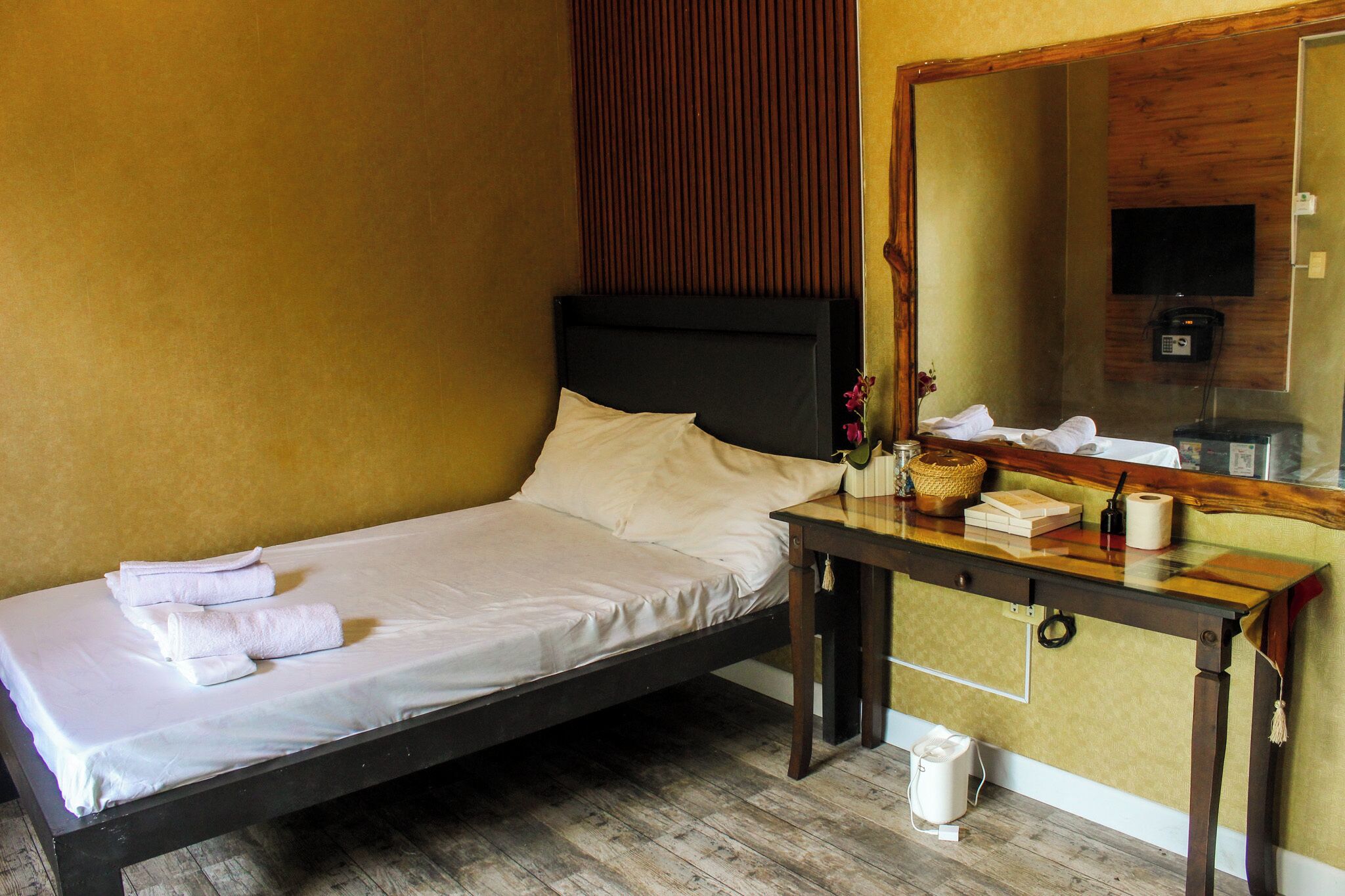 Un lit simple avec du linge de lit et des serviettes blanches, une table de chevet avec des articles de toilette, un miroir et un plancher en bois.