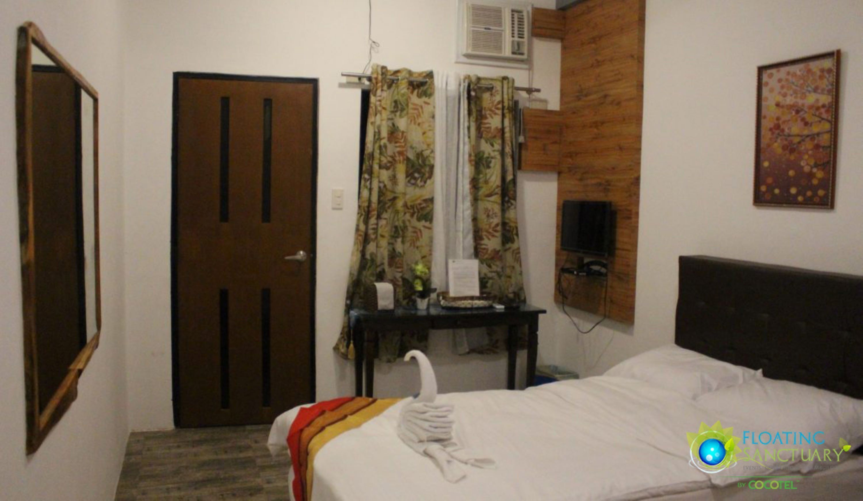 A hotel room with a bed, a wooden headboard, a TV mounted on a wooden panel, a desk with a chair, a mirror, and a door.