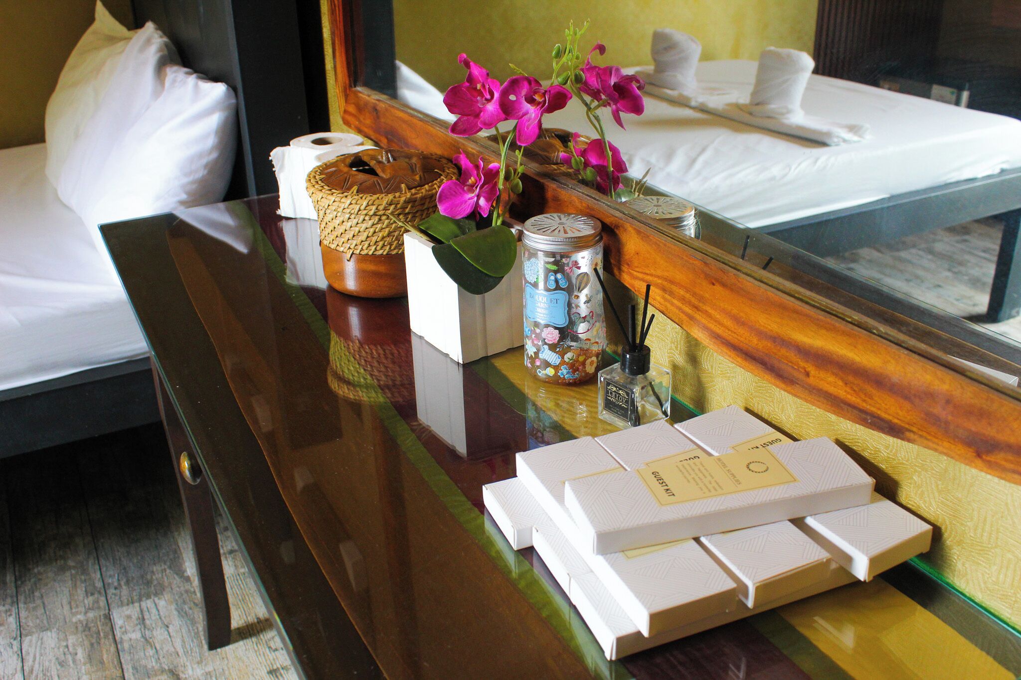 Une chambre d’hôtel avec un lit bien fait, une table de chevet surmontée d’un panier en osier, un bocal étiqueté et un miroir qui reflète le lit et les oreillers.