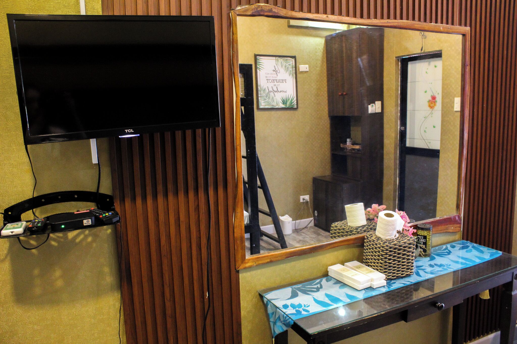 A room with a flat-screen TV mounted on the wall, a wooden cabinet, a mirror, and a table with toiletries.