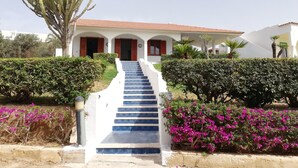 Un edificio con una scalinata che conduce a un ingresso con tetto in tegole rosse, circondato da siepi e piante fiorite.