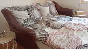 Sängyssä on kuviollinen päiväpeite, ruskea nahkapääty ja pieni yöpöytä, jossa on paperinenäliöpakkaus.